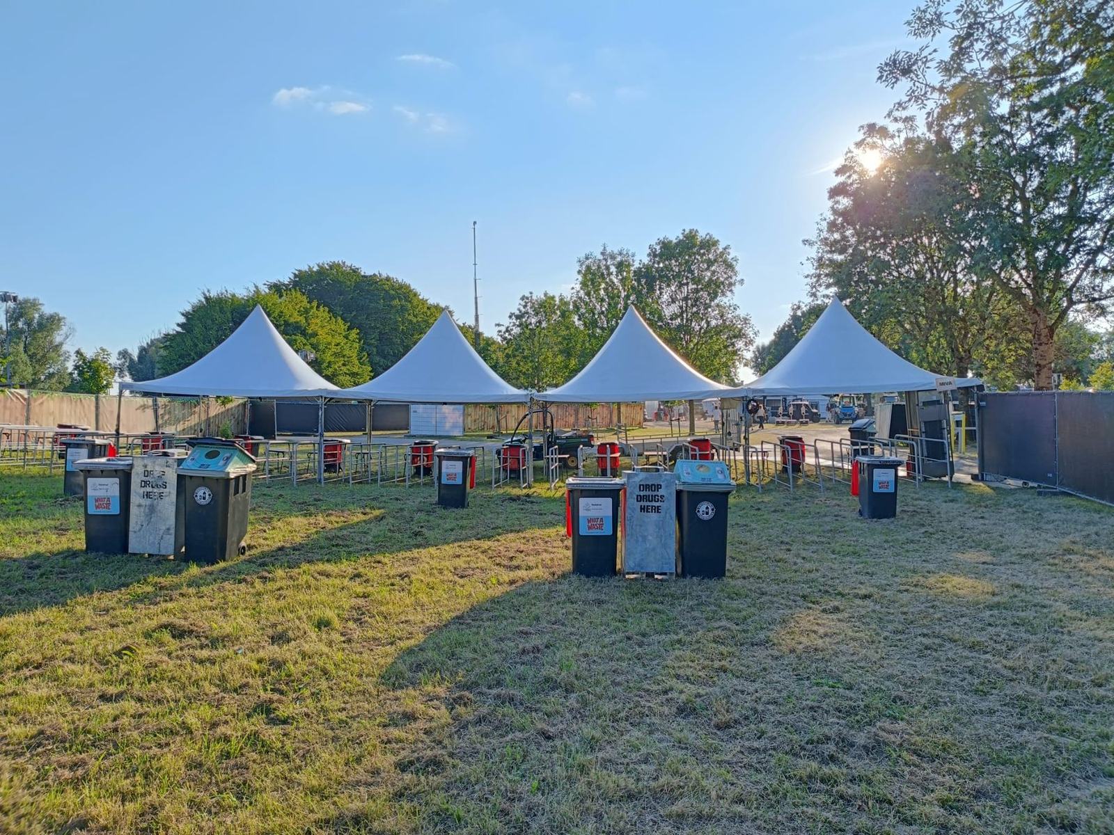 Inzamelpunten met rolcontainers bij de ingang van een festivalterrein, met tenten op de achtergrond