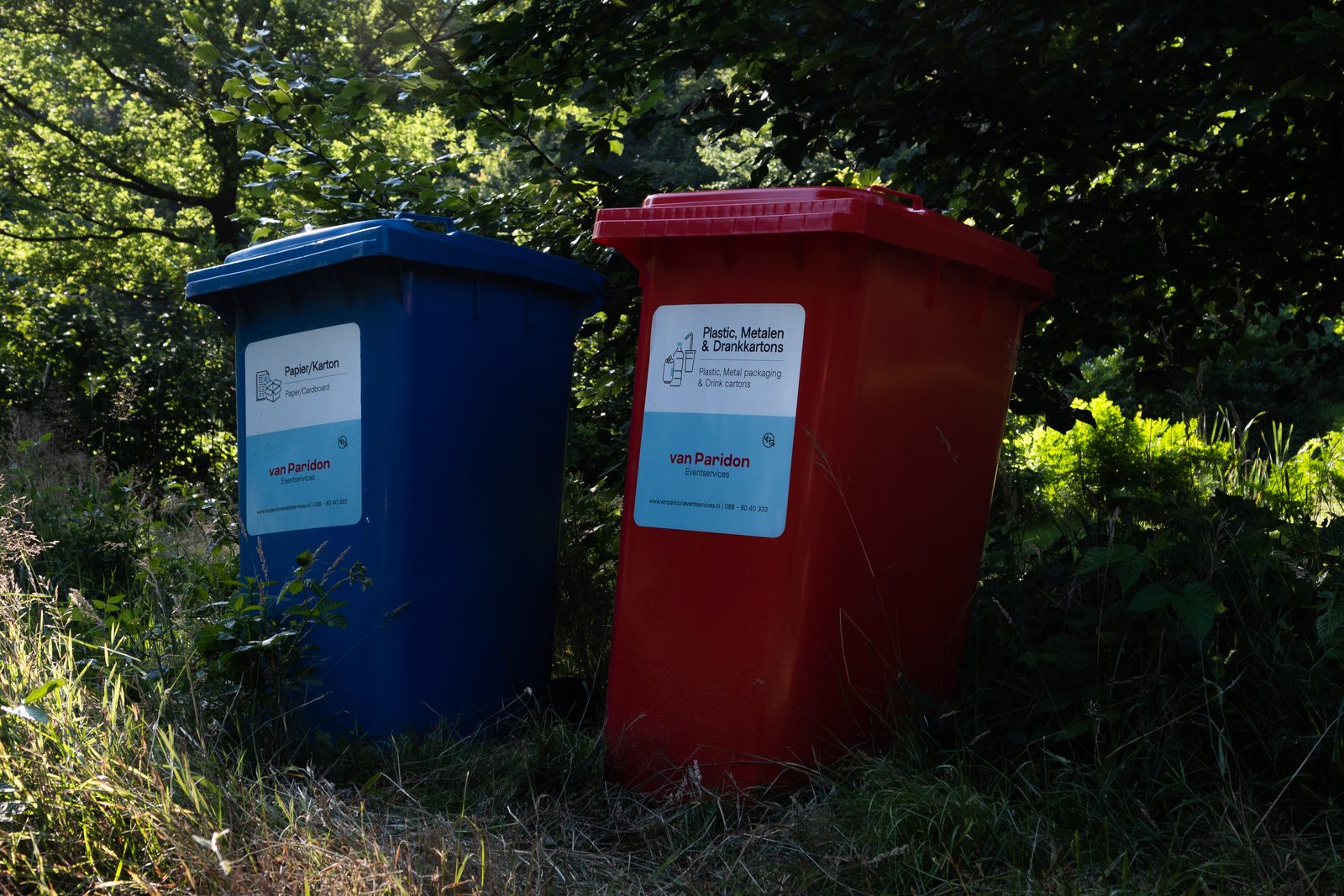Een blauwe en een rode rolcontainer met stroomlabels tussen het groen op festivalterrein Wonderfeel